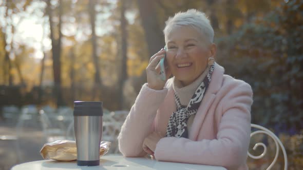 Portrait of Beautiful Aged Woman in Stylish Clothes Talking on Smartphone Sitting at Table in Park