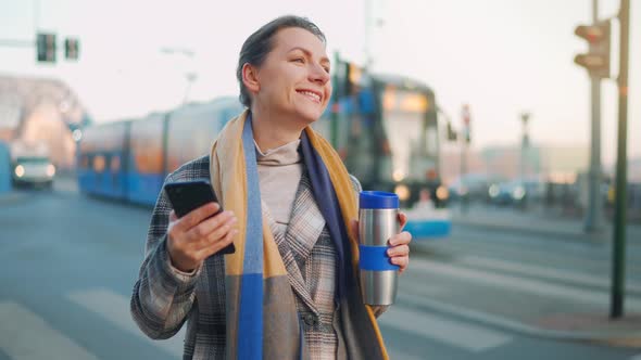 Woman in a Coat Standing on the Street in the Early Morning and Using Smartphone