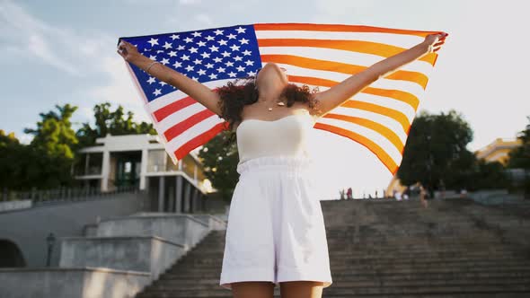 Darkskinned Model in White Outfit is Smiling Raising Flag of USA Above Her Head and Waving It While