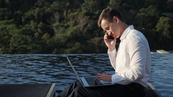 Man Talks on Phone Typing on Laptop Against Forest and Pool