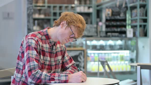 Focused Young Redhead Man Writing on Paper in Cafe 