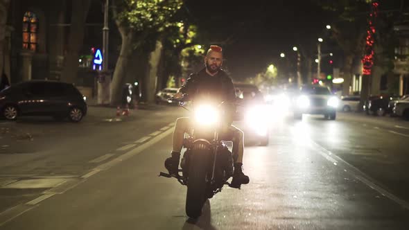 Front View of a Brutal Motorcyclist Drives a Bike in the City with Bright Headlight at Night