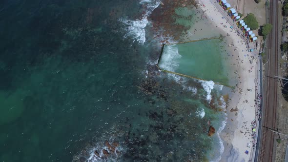 Top down shot from drone of train tracks next to ocean with tide pool, coloured beach shacks on beac