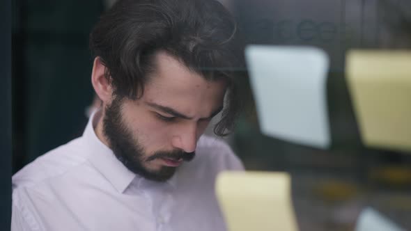 Closeup Thoughtful Bearded Man Standing at Glass Door with Adhesive Notes Analyzing Business