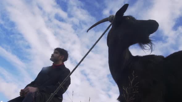 A Young Stylish Guy Walks with a Goat Near His House in Forest