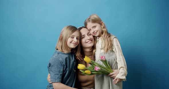 Mom Hugs Daughters and Kisses with Flowers Posing on a Blue Background