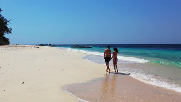 Man and woman holding hands, walking on wet sand of exotic beach, watching white waves of blue turqu