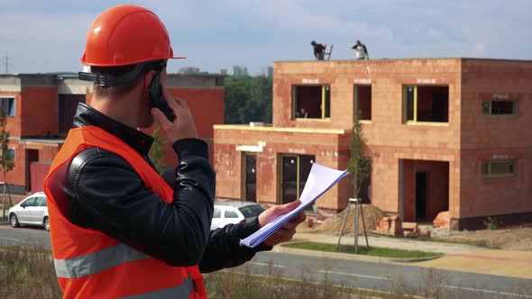 A Young Construction Worker Talks on a Smartphone and Looks at Papers