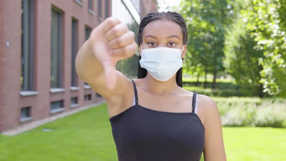 A Young Black Woman in a Face Mask Shows a Thumb Down To the Camera and Shakes Her Head