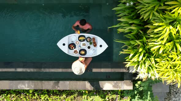 Aerial top down view of young couple with a floating breakfast poolside at a tropical luxury villa d