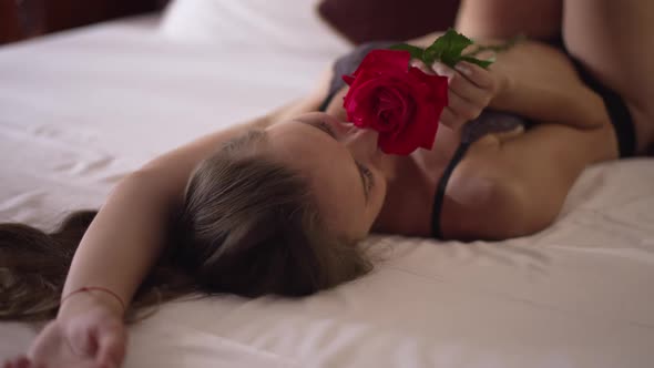 Young Slim Woman Lying on Comfortable Bed in Lingerie Smelling Red Rose in Slow Motion Smiling
