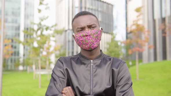 A Young Black Man in a Face Mask Thinks About Something - Office Buildings in the Blurry Background