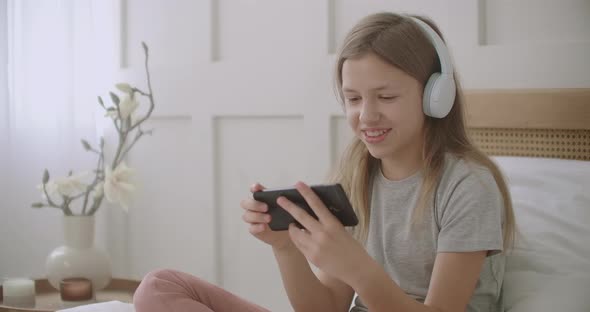 Little Girl Is Using Smartphone and Wireless Headphones Sitting on Bed in Room, Playing Game and