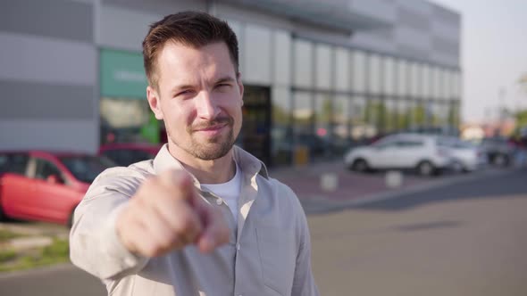 A Young Caucasian Man Points at the Camera and Nods in an Urban Area