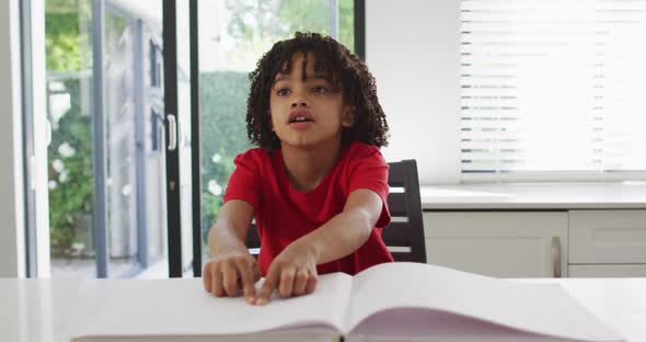 Happy biracial boy sitting at table in kitchen reading braille