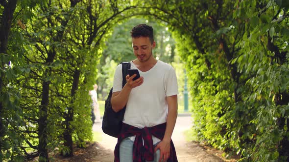 Handsome Young Man Stands in the Park Inside a Tunnel of Trees with a Phone in His Hands