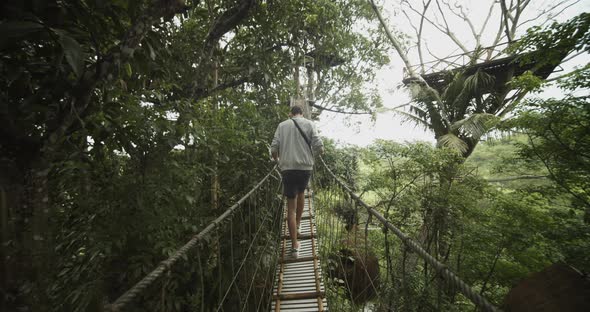 Dolly View Following a Man Carefully Walking Across a Shaking Hanging Bridge in a Tropical