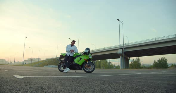 A Man Stands By a Motorcycle and Looks at the Time