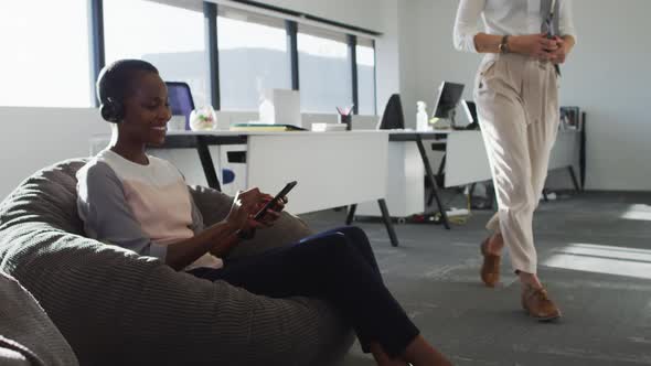 African american businesswoman wearing headphones, sitting in armchair while her coworker passing by