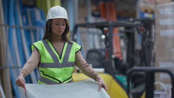 Expert Female Contractor Examining Blueprint Standing in Warehouse in Hard Hat and Vest