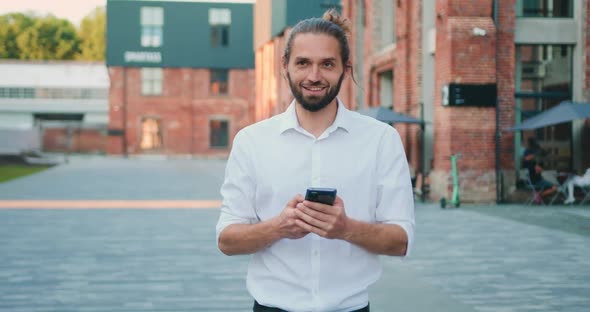 Smiling Man in a White Shirt Type Message on Phone Walking Outdoor.