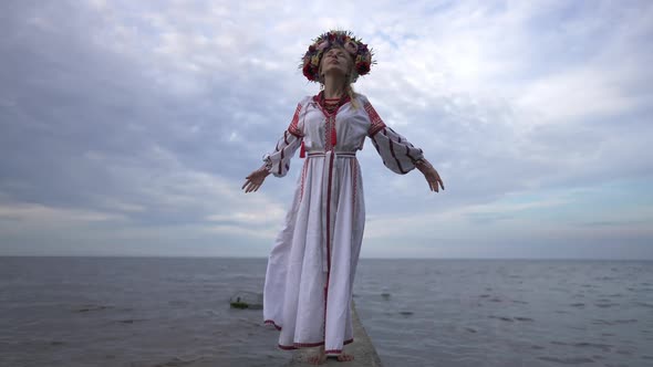 Young Calm Confident Ukrainian Woman in Embroidered Dress Standing on River Pier Raising Hands in