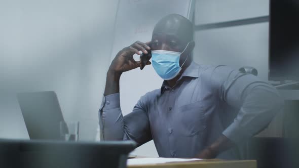 African american businessman in face mask talking on smartphone, using laptop in conference room