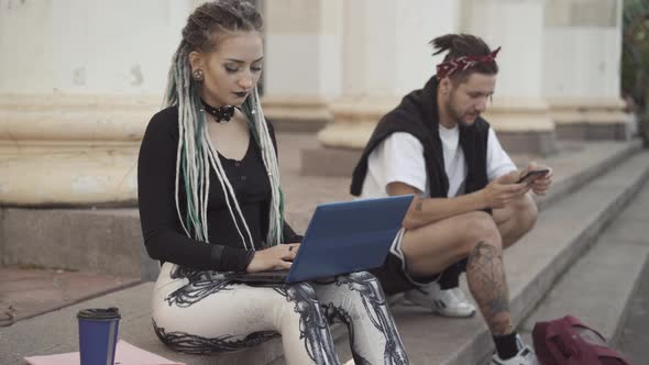 Portrait of Concentrated Goth Woman Messaging Online on Laptop with Blurred Man Using Smartphone and