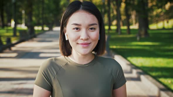 Outdoor Portrait of Young Asian Woman Listening to Music in Airpods and Smiling to Camera in Park