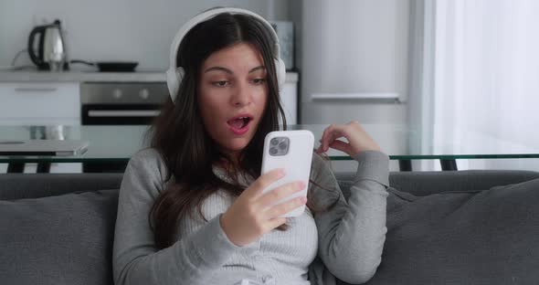 A Shocked Brunette Girl in a Grey Sweater Saying Wow Looking Smartphone and Covering Her Mouth While