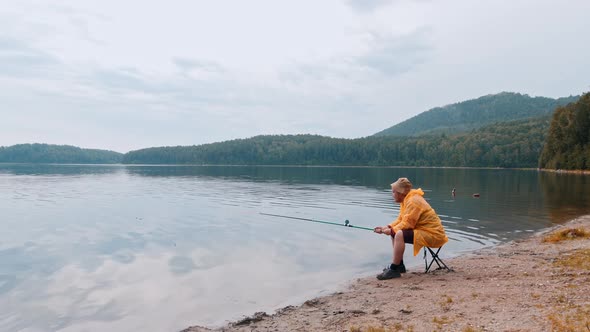 Lonely Man in a Raincoat Sitting on a Shore and Fishing