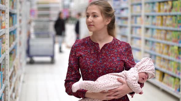 Woman Choosing Children Food with Little Baby Child Girl on Hands During Supermarket Shopping