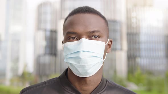 A Young Black Man in a Face Mask Looks Around - Face Closeup - Office Buildings in the Background