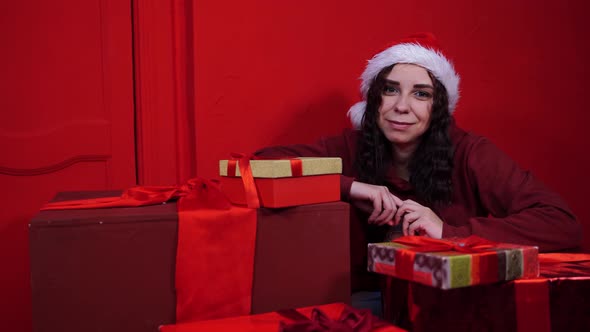 Young Woman in Santa Claus Hat Sitting in Gifts Near Red Wall