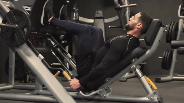 Side View of Young Man Exercising on Leg Press Machine in Gym 