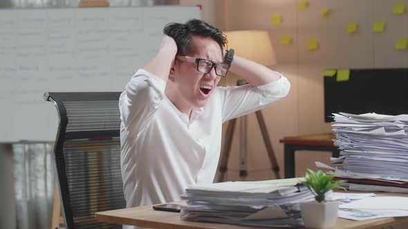 Asian Man Having Headache While Working Hard With Documents At The Office