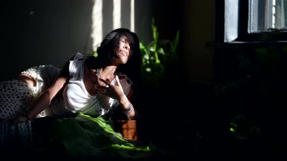 Sexy Woman is Lying on Couch in Sunny Morning in Beautiful Room with Plants Resting in Greenhouse