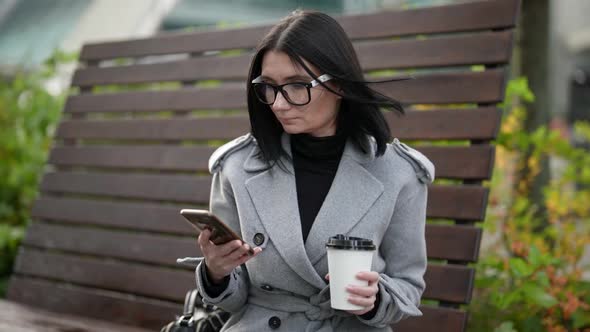 Businesswoman is Checking Email By Smartphone Sitting in Park in Morning Holding Cup with Coffee