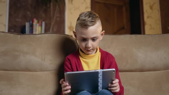 Small Boy in Homewear Which Sitting on Comfortable Couch at Home and Uses Tablet Device