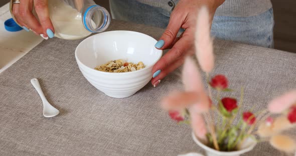 Female Hand Holding Bottle Pouring Milk in Cereal Granola Flakes Bowl