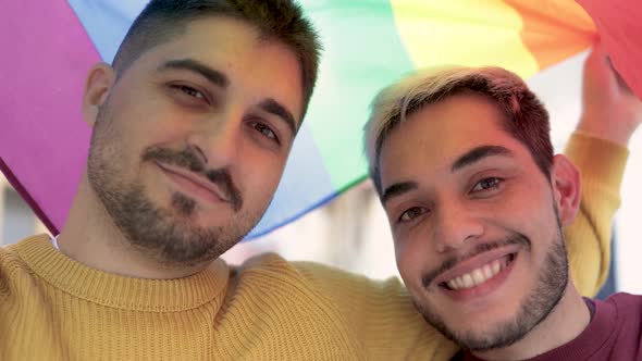 Young Gay Men Couple Having Tender Moment Under Rainbow LGBT Flag Outdoor  LGBTQ Love Concept