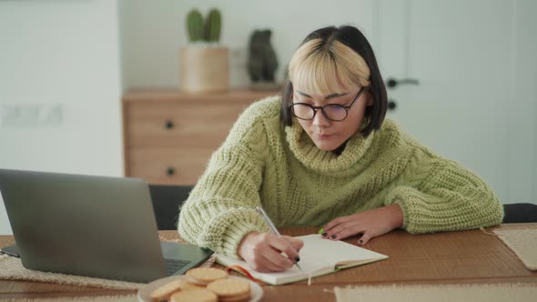 Serious Asian woman in eyeglasses writing something in notepad from laptop