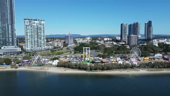 Oceanside carnival set-up along a coastal beach strip below a towering city landscape. Fast panning