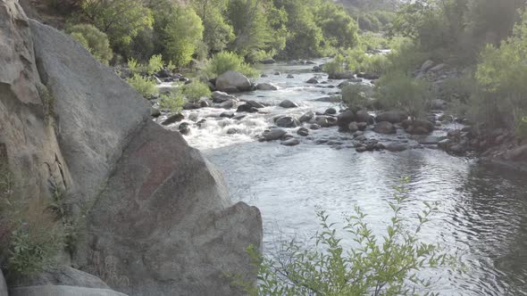 A drone flies out from behind a set of rocks revealing a tranquil stream and river.