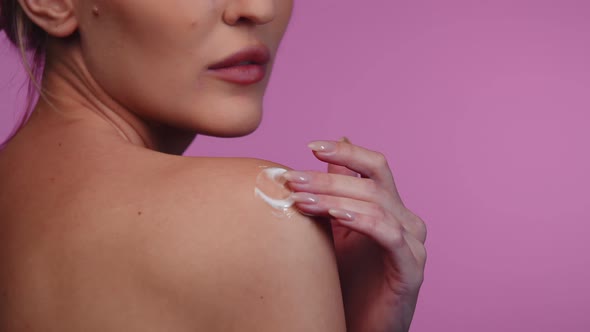 Woman Rubs Dermatological Cream with Vitamins Into the Skin on Shoulder Pink Background