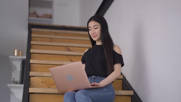 Slim Gorgeous Concentrated Asian Woman Typing on Laptop Keyboard Sitting in Home Office on Stairs