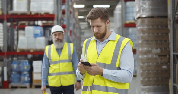 Aged Warehouse Manager Giving Helmet to Young Worker Standing in Using Smartphone