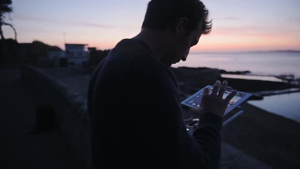 Man piloting a drone with a controller, above outdoor swimming pools, Guernsey seaside, at dusk