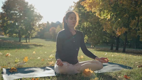 Young Woman Sitting on Mat in Lotus Position in City Park on Sunny Autumn Day