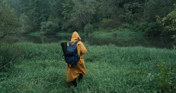 Hiker in a Yellow Raincoat with a Backpack in the Tall Grass Next to the River